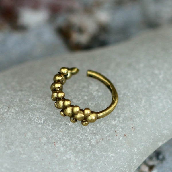 A close-up of the ROE Beaded Nose Ring in Gold by Kaya Movement, a small 20 gauge gold-tone brass piece featuring an intricate beaded tribal ethnic design. The textured round beads along its curved band add charm as it rests on a smooth, light-colored stone surface, with a blurred background enhancing its elegance.